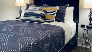 Une chambre moderne avec un grand lit, une tête de lit sombre et un tableau au-dessus du lit. Il y a deux tables de chevet avec des lampes.