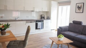 Eine moderne Küche mit weißen Schränken, eine Essecke mit einem Holztisch und einem grau gepolsterten Stuhl, und ein graues Sofa in einem Wohnzimmer.