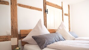 Ein ordentlich gemachtes Bett mit Holzkopfende, ein Nachttisch mit Lampe und Blumenvase, und eine weiße Tür im Hintergrund.