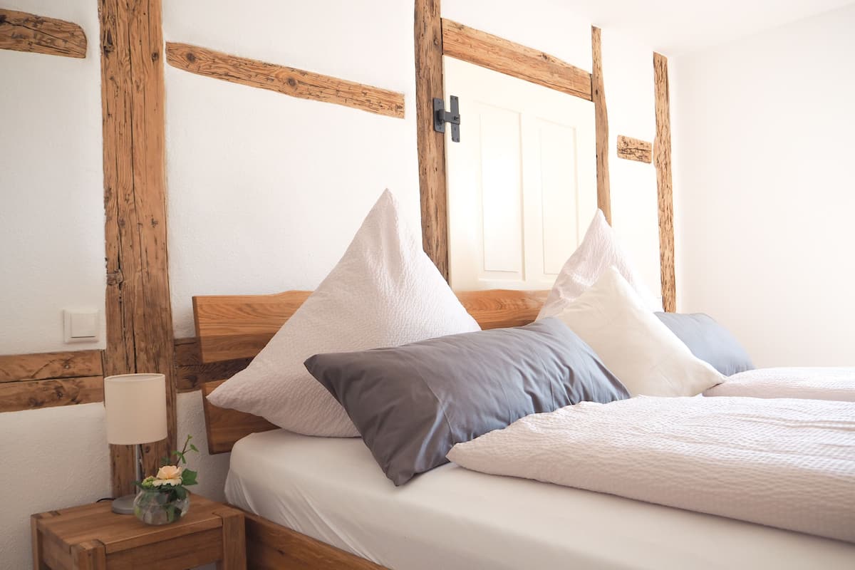 A neatly made bed with a wooden headboard, a bedside table with a lamp and a vase of flowers, and a white door in the background.