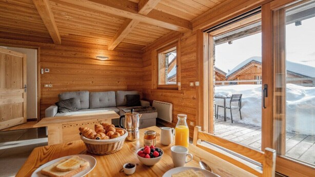 A wooden cabin interior with a dining table set for breakfast, a sofa, and a view of a snowy landscape through a glass door.