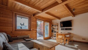 A cozy cabin interior with a wooden ceiling, a sofa, a dining table, and a TV mounted on the wall.
