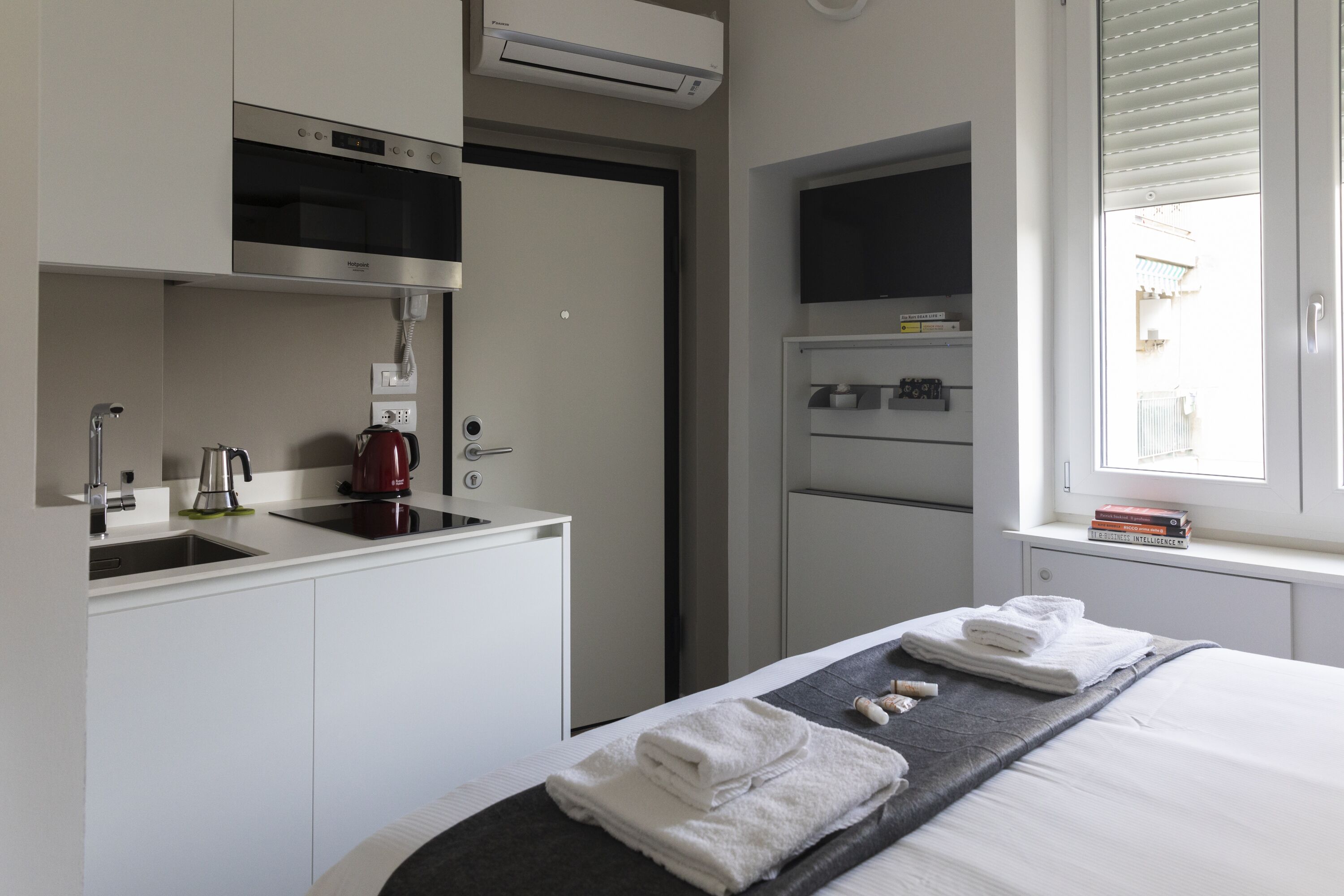 A compact hotel room with a kitchenette, a bed with neatly folded towels, a window with blinds, and a mounted television.