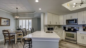 A modern kitchen with white cabinetry, a large island, stainless steel appliances, and a dining area with wooden chairs and a table. - 5105 Hampton Place - Amazing Views (Hilton Head Island)