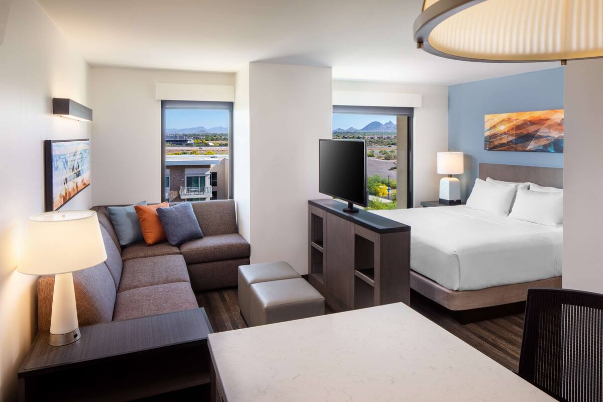 Habitación de hotel con cama, sofá, escritorio, televisor y vista a la ciudad.