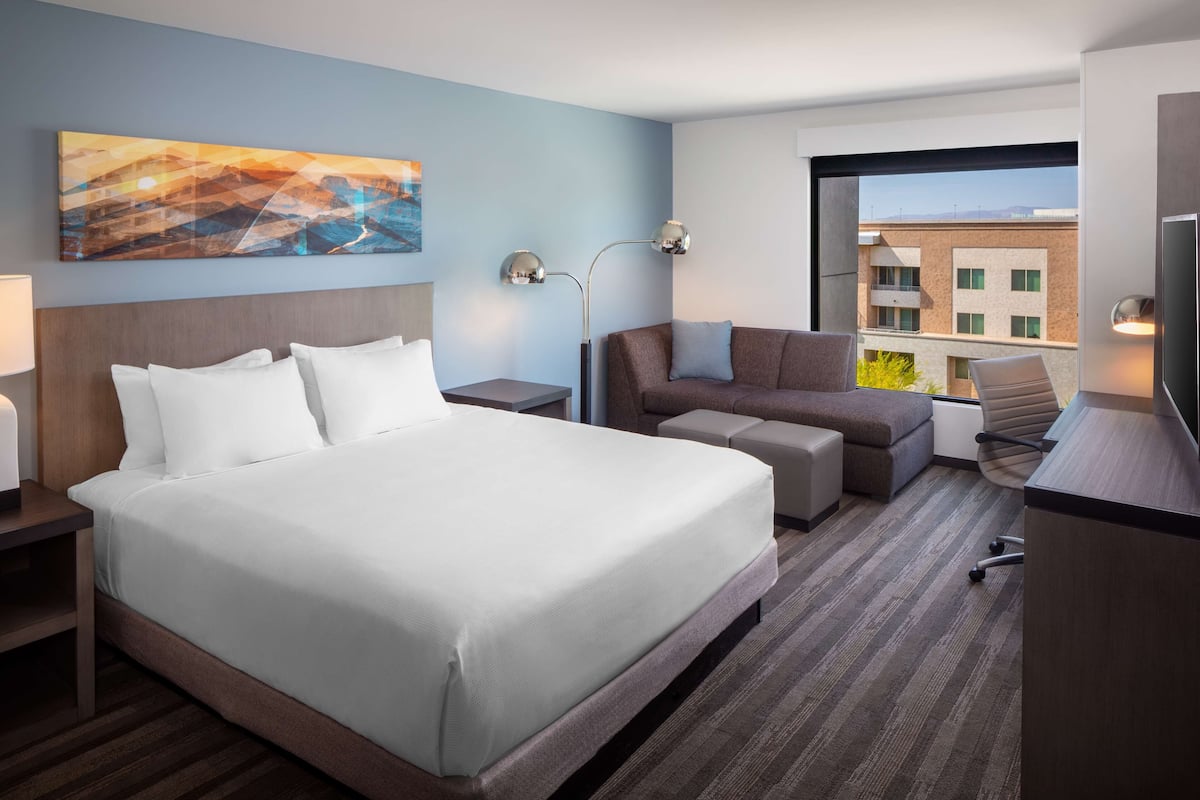 Habitación de hotel con una cama grande, un sofá, un escritorio y un televisor. Hay un cuadro en la pared y una ventana con vista a edificios.