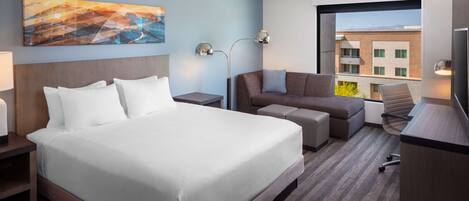 A hotel room with a large bed, a sofa, a desk, and a TV. There is a painting on the wall and a window with a view of buildings.