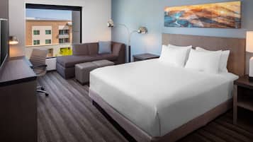A hotel room with a large bed, a sofa, a desk, and a chair. There is a window with a view of buildings outside.