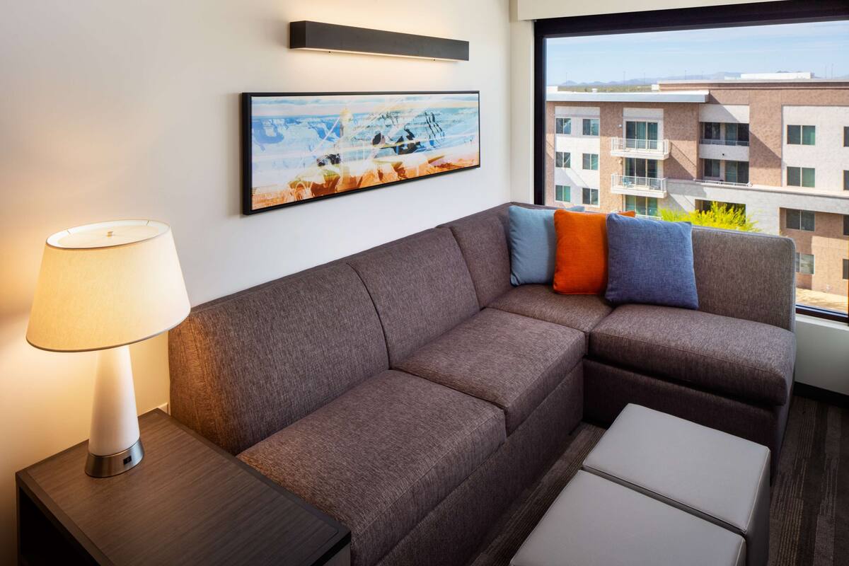 Una habitación de hotel moderna con sofá modular, una mesita de noche con lámpara y un ventanal con vistas a la ciudad.