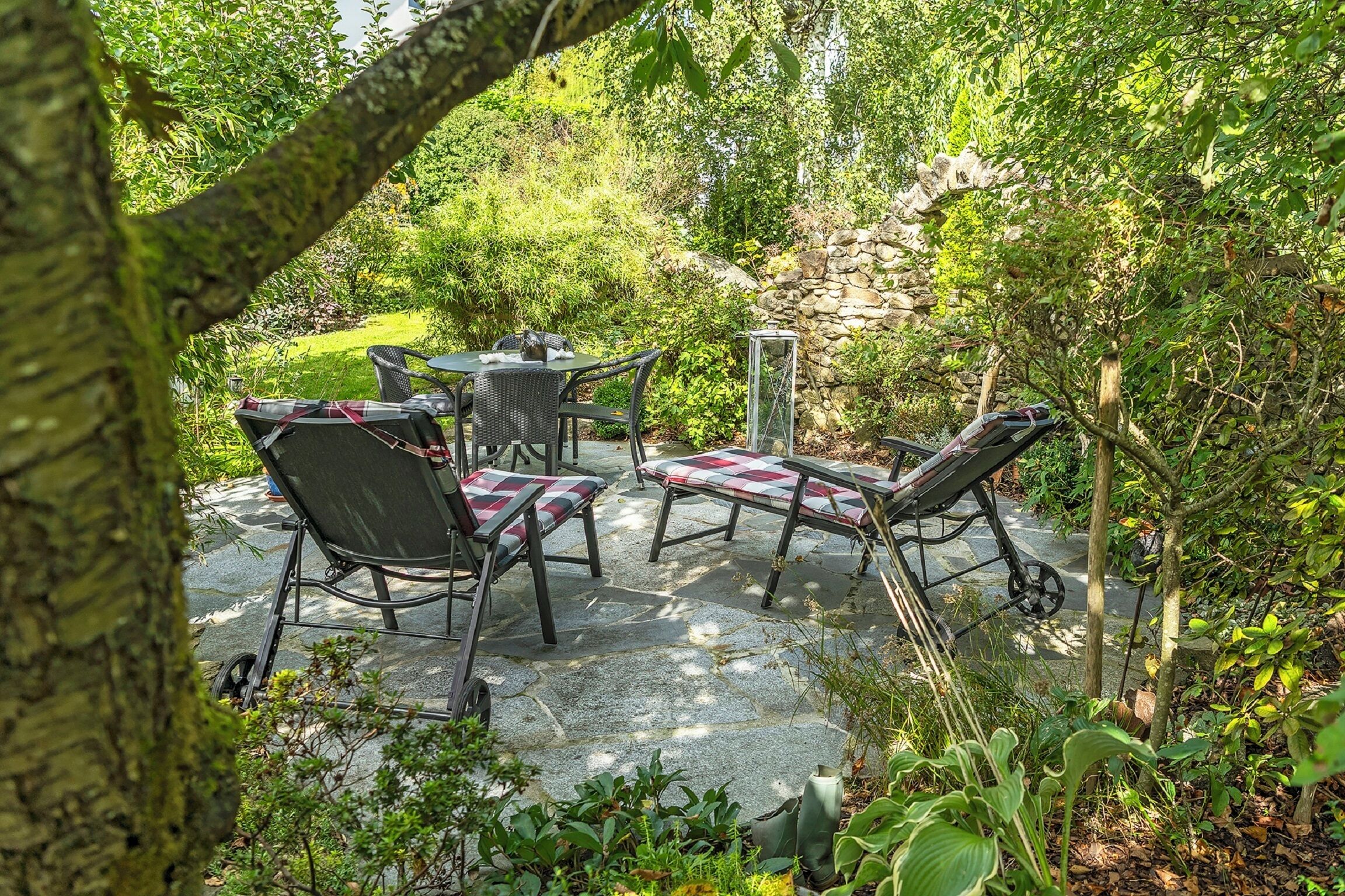Un patio exterior con una mesa y sillas, rodeado de abundante vegetación y un muro de piedra.