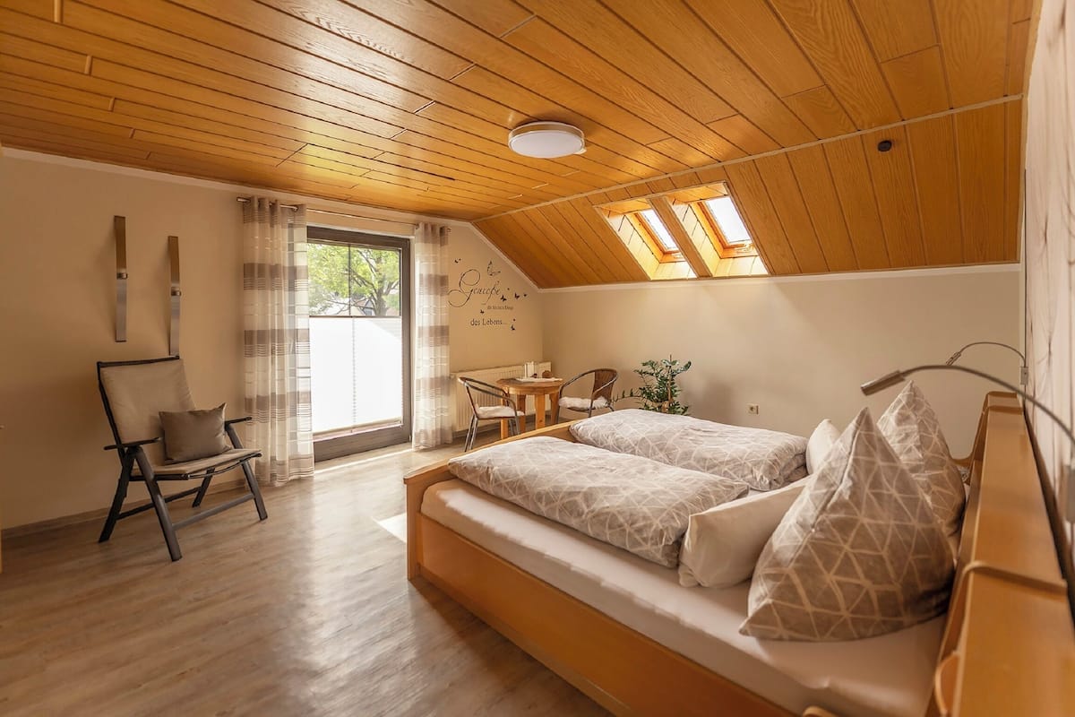 A bedroom with a wooden ceiling, two beds, a chair, a table, and a window with curtains.