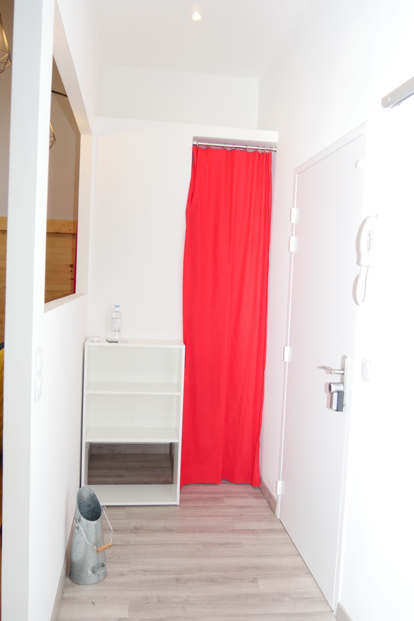 A narrow hallway with a white door, a red curtain, a white shelf unit, and a metal bucket.