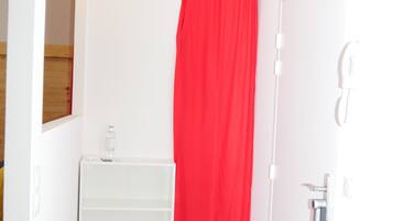 A narrow hallway with a white door, a red curtain, a white shelf unit, and a metal bucket.