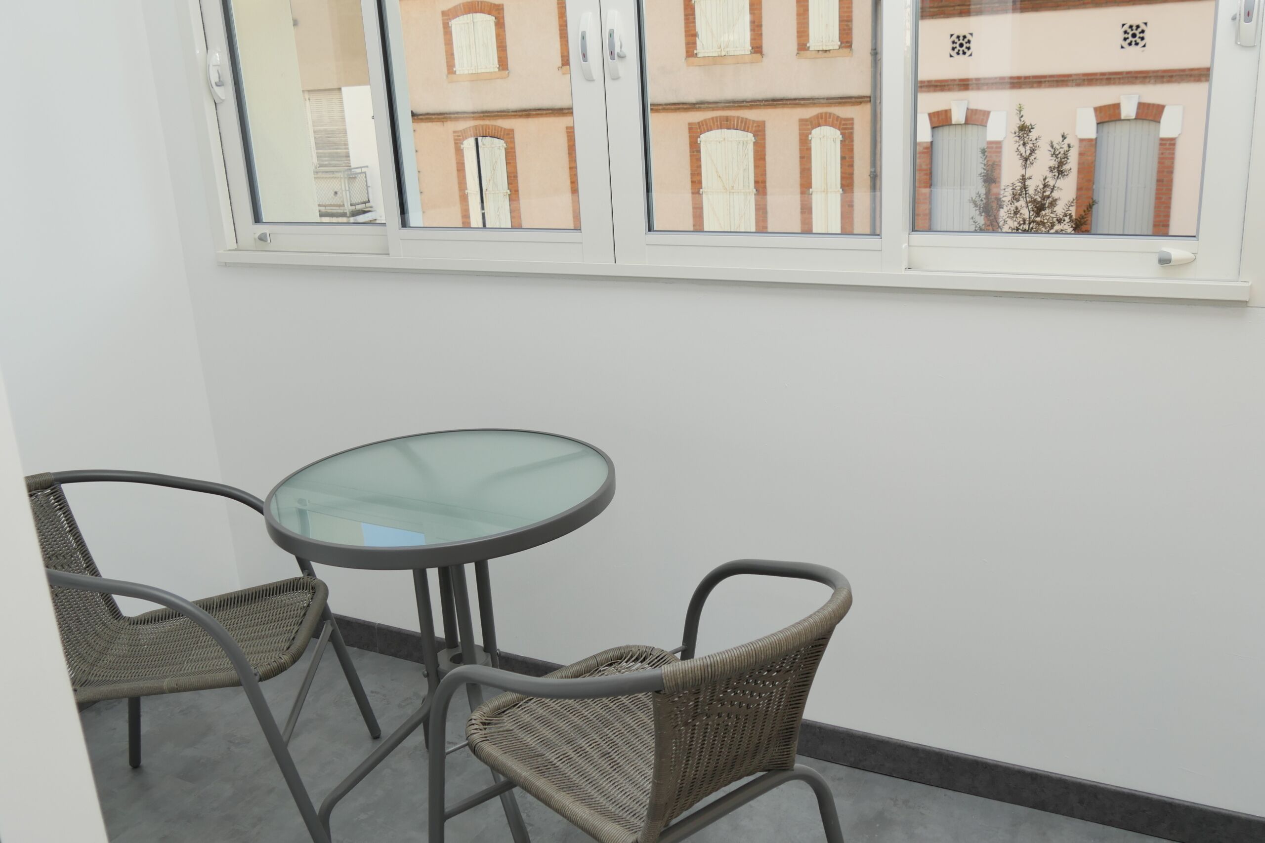 A balcony with a glass table and wicker chairs, overlooking a building with arched windows.