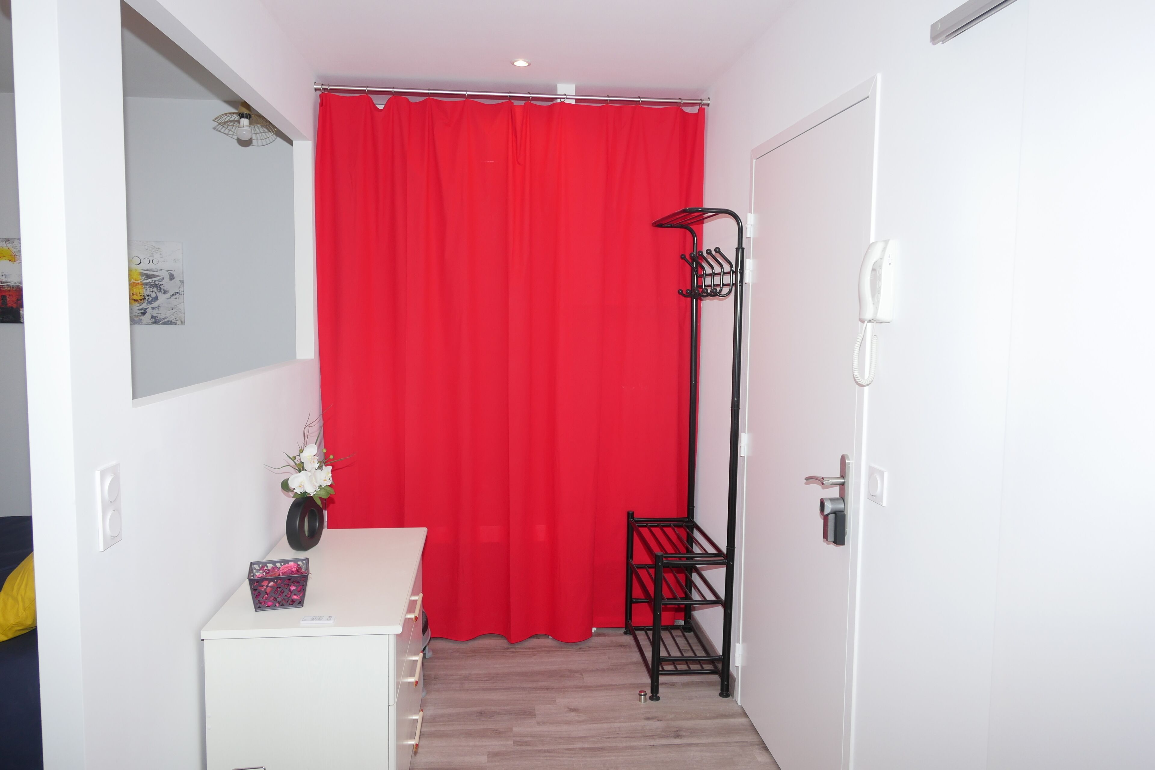A small room with a red curtain, a white desk with drawers, a vase with flowers, and a black metal rack.