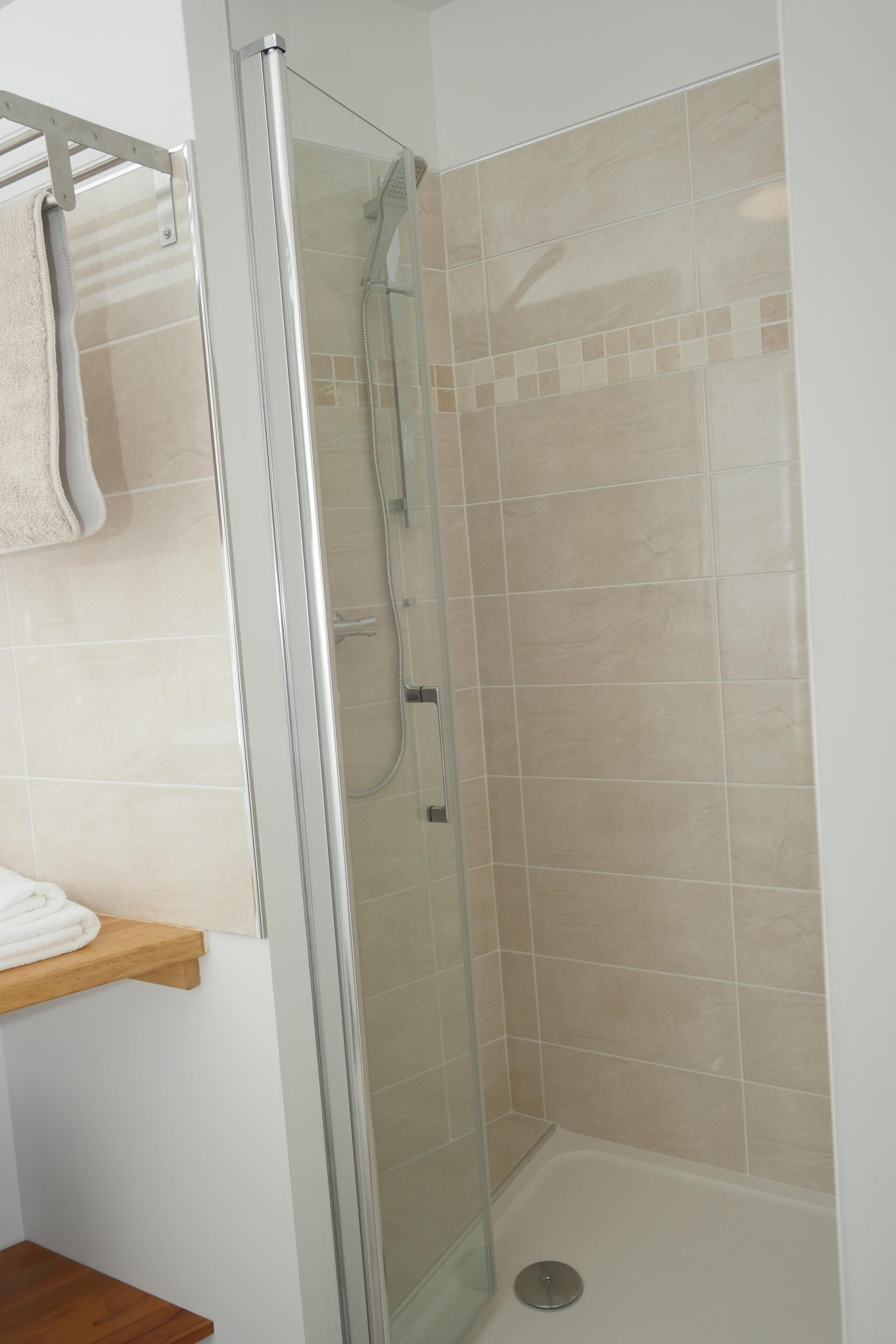 Una ducha con cerramiento de vidrio, azulejos color beige, un estante de madera con una toalla y piso de madera.