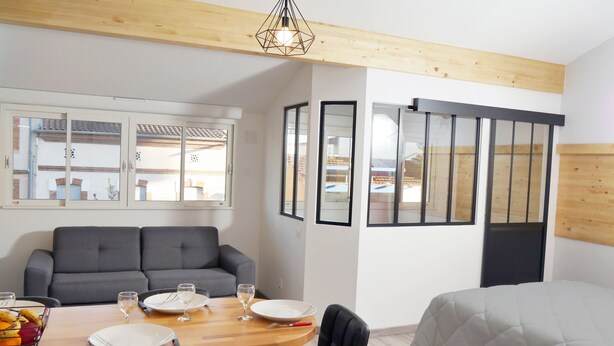 A modern room with a dining table set for two, a grey sofa, a bed with a white duvet, and large windows with a view of the outside. - Appart Hôtel Albi (Albi)