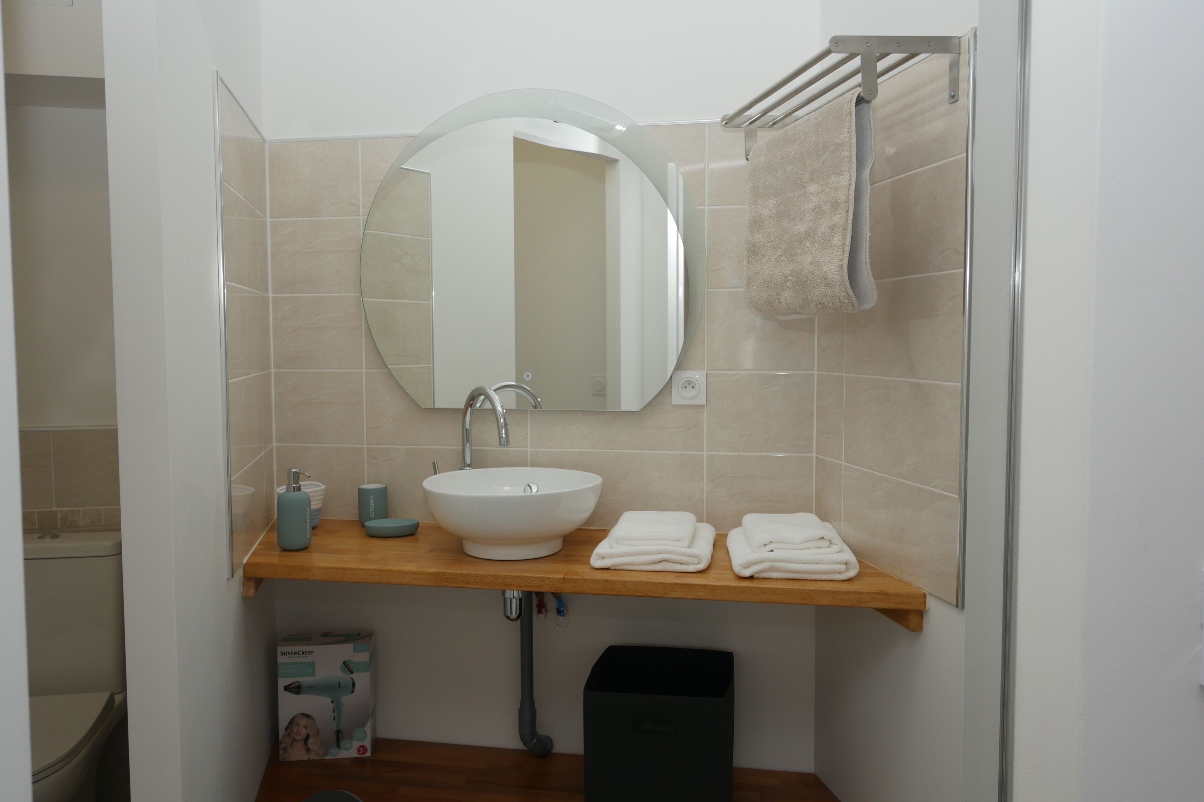 Une salle de bain avec un miroir rond, un lavabo blanc, un plan de toilette en bois et un porte-serviettes.