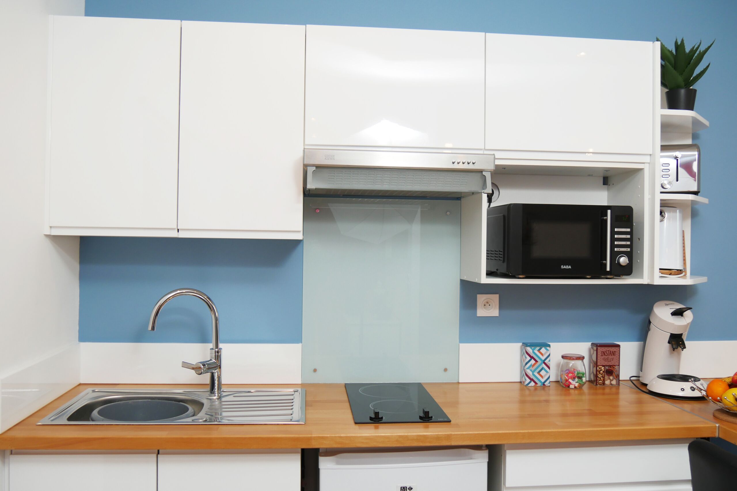 A compact kitchen with white cabinets, a stainless steel sink, a microwave, a coffee maker, and a small plant on the counter.