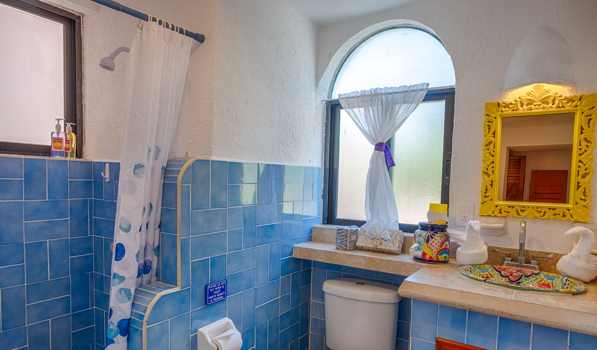 A bathroom with blue tiles, a shower curtain, a toilet, a sink with a decorative mirror, and a window with a curtain.