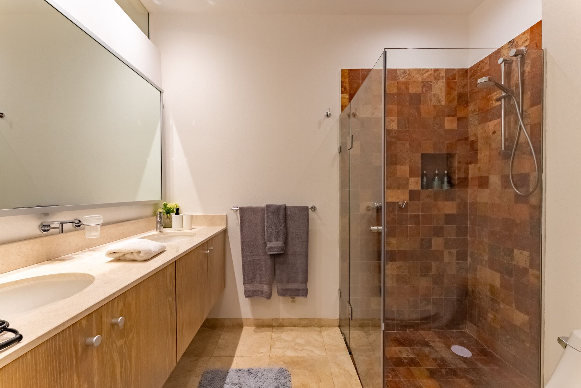 Un bagno moderno con doccia vetrata, un ampio specchio, mobili contenitori in legno e un pavimento piastrellato color beige.