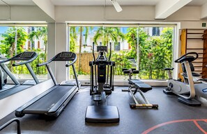 A gym with treadmills, an elliptical, and weight machines, with large windows offering a view of palm trees and buildings.