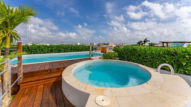 A rooftop swimming pool with a wooden deck and a hot tub, surrounded by greenery and overlooking a cityscape. - Downtown Playa close 5th Av Rooftop Pool (Playa del Carmen)