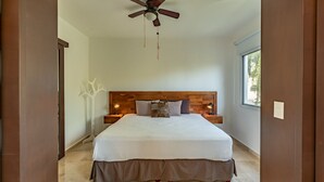 A bedroom with a wooden headboard, a bed with a white and brown bedspread, two bedside lamps, a ceiling fan, and a window with a view of greenery. - Downtown Playa close 5th Av Rooftop Pool (Playa del Carmen)