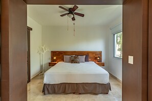 Ein Schlafzimmer mit einem hölzernen Kopfteil, einem Bett mit weißer und brauner Bettdecke, zwei Nachttischlampen, einem Deckenventilator und einem Fenster mit Blick ins Grüne.