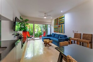 A modern living room with a blue sofa, a dining table, wicker chairs, a TV, and a large window with a view of greenery.