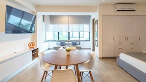 Un salon moderne avec une table ronde en bois, des chaises blanches, un canapé gris agrémenté de coussins à motifs et un téléviseur fixé au mur.