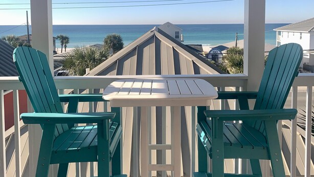 Un balcon avec deux fauteuils Adirondack verts et une petite table blanche, donnant sur une plage et l’océan.