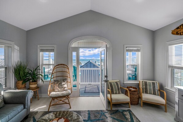 A spacious living room with a high ceiling, a large window, and a view of the beach. There are two wicker chairs, a sofa, and a small table with plants.