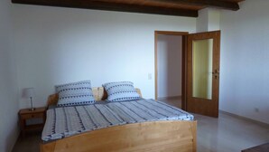 Quarto com cama de madeira, duas almofadas, mesa de cabeceira com abajur e porta que dá para outro cômodo.