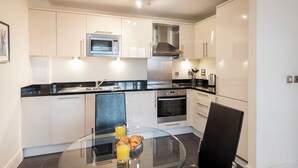 A modern kitchen with white cabinets, a black countertop, a stainless steel microwave, and an oven. There is a glass table with a plate of croissants and a glass of orange juice. - One Bed Apartment in Waterloo near Southwark (London)