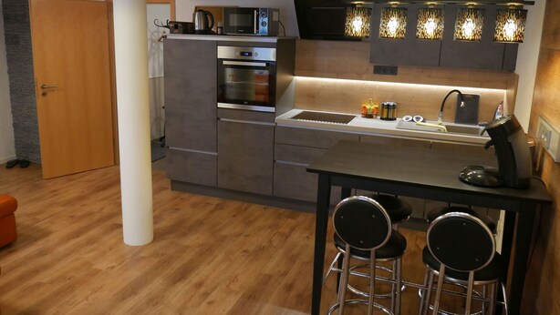 A compact kitchen with a microwave, oven, and modern lighting. There are two bar stools and a wooden door. - Haus Alexandra (Schwarzenbach an der Saale)
