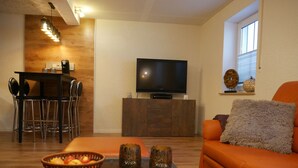 A living room with a sofa, a coffee table with fruit and remotes, a TV on a wooden cabinet, and a dining area with chairs and a table. - Haus Alexandra (Schwarzenbach an der Saale)