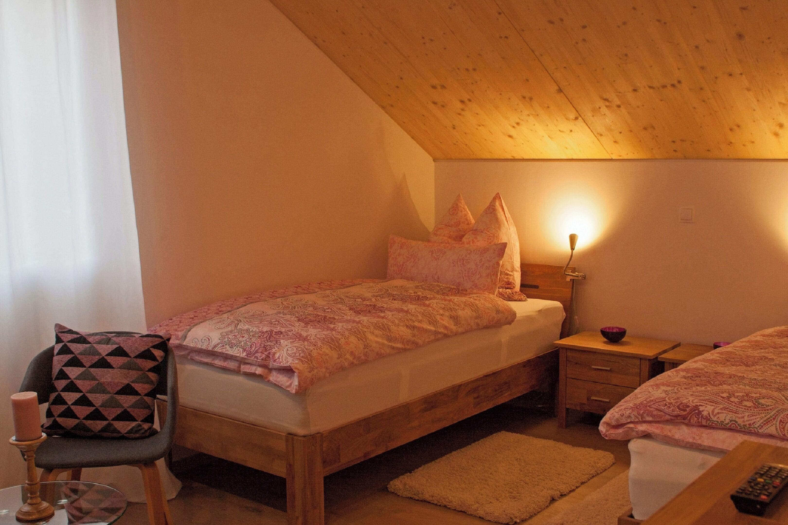 Ein gemütliches Schlafzimmer mit Holzboden, einem Bett mit gemusterten Bettwäsche, einem Nachttisch mit Lampe und einem Sessel mit geometrischem Kissen.