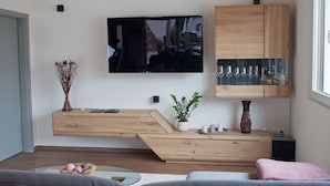 Un salon moderne avec un téléviseur à écran plat fixé au mur, des meubles en bois, une vitrine en verre contenant des verres à vin et un coin salon confortable avec un canapé et des coussins.