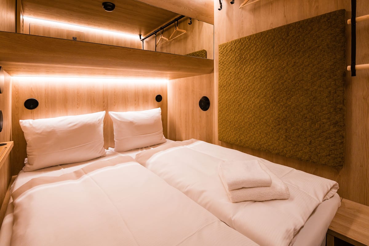 Una cama bien hecha con sábanas blancas y una toalla doblada, cabecera de madera y un panel de pared texturizado.