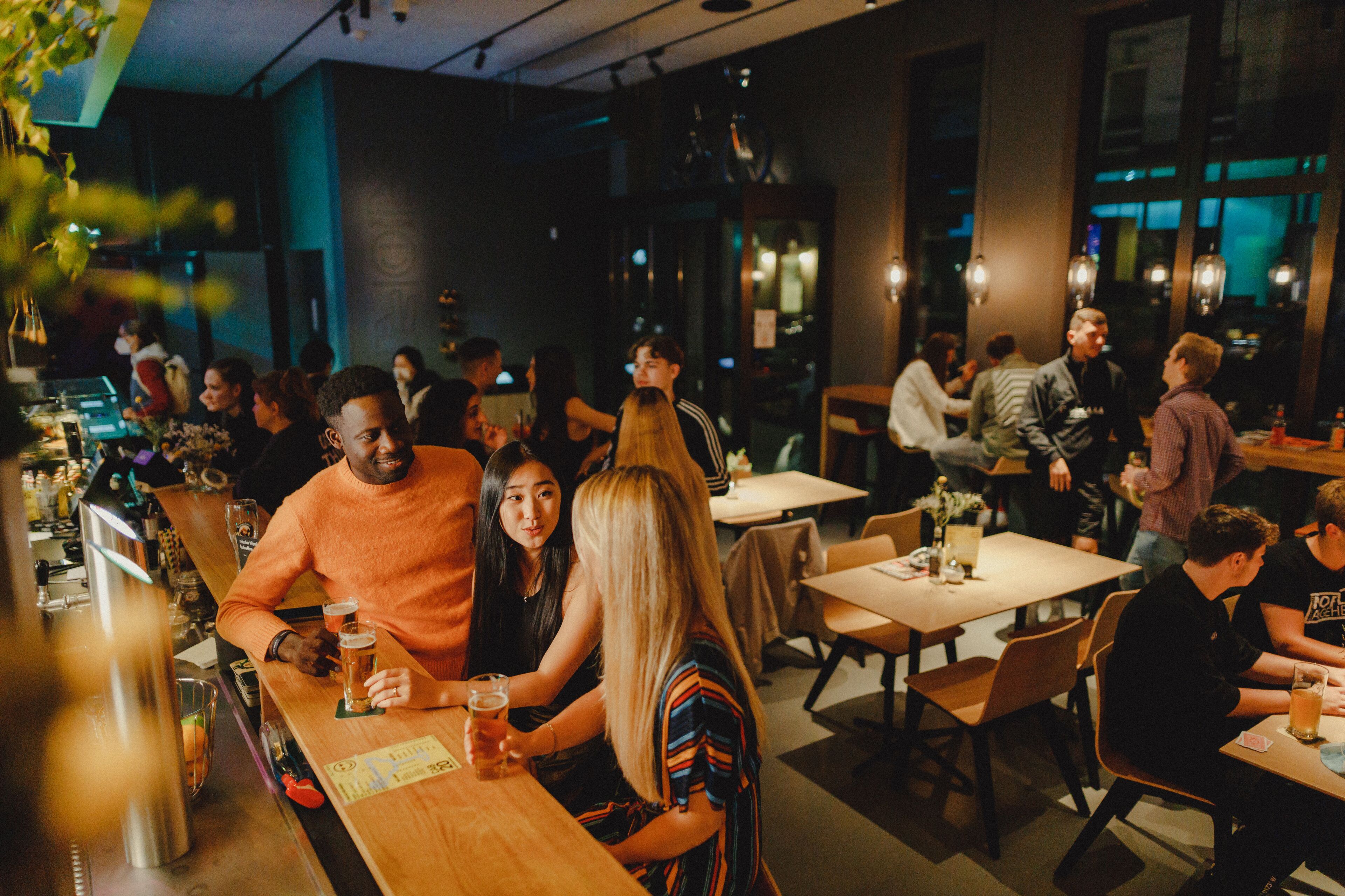 A bar area with tables and chairs, people sitting and standing, and a bartender at the counter.