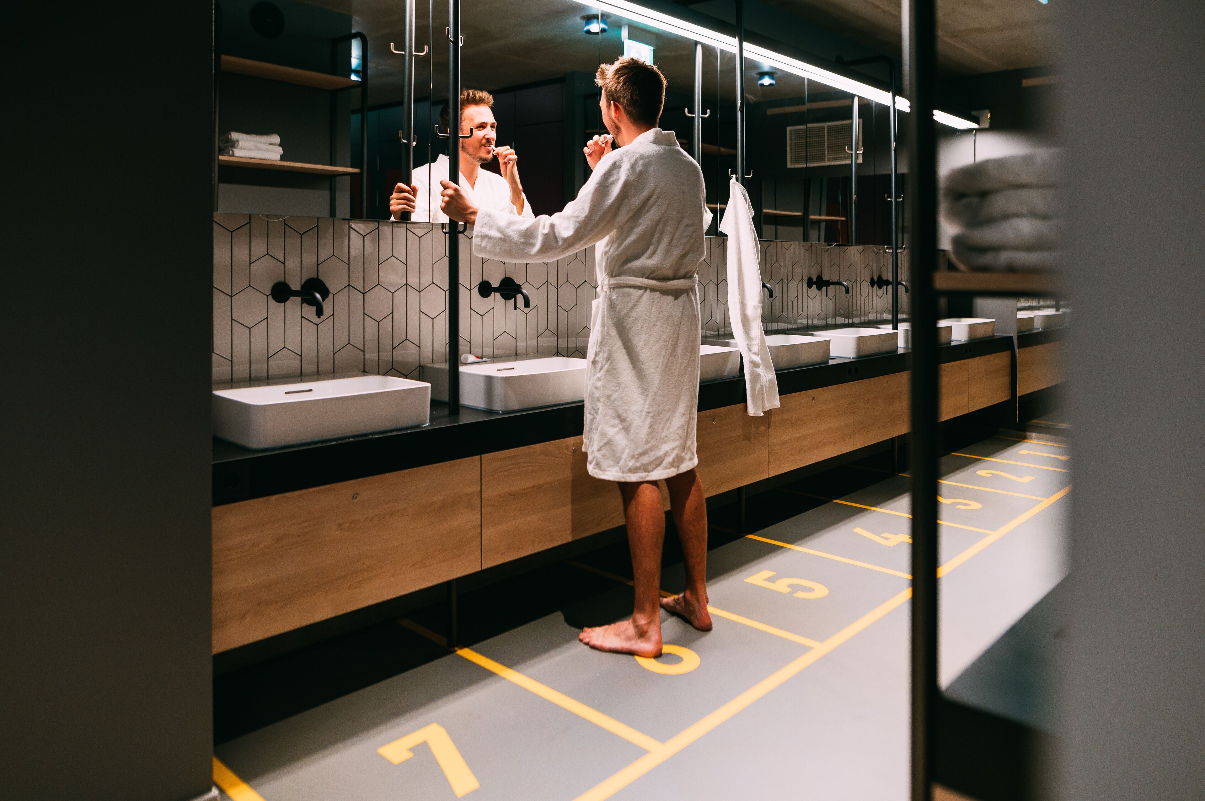 Una persona con bata de baño en un baño moderno con un espejo grande, varios lavamanos y un revestimiento de azulejos geométrico.
