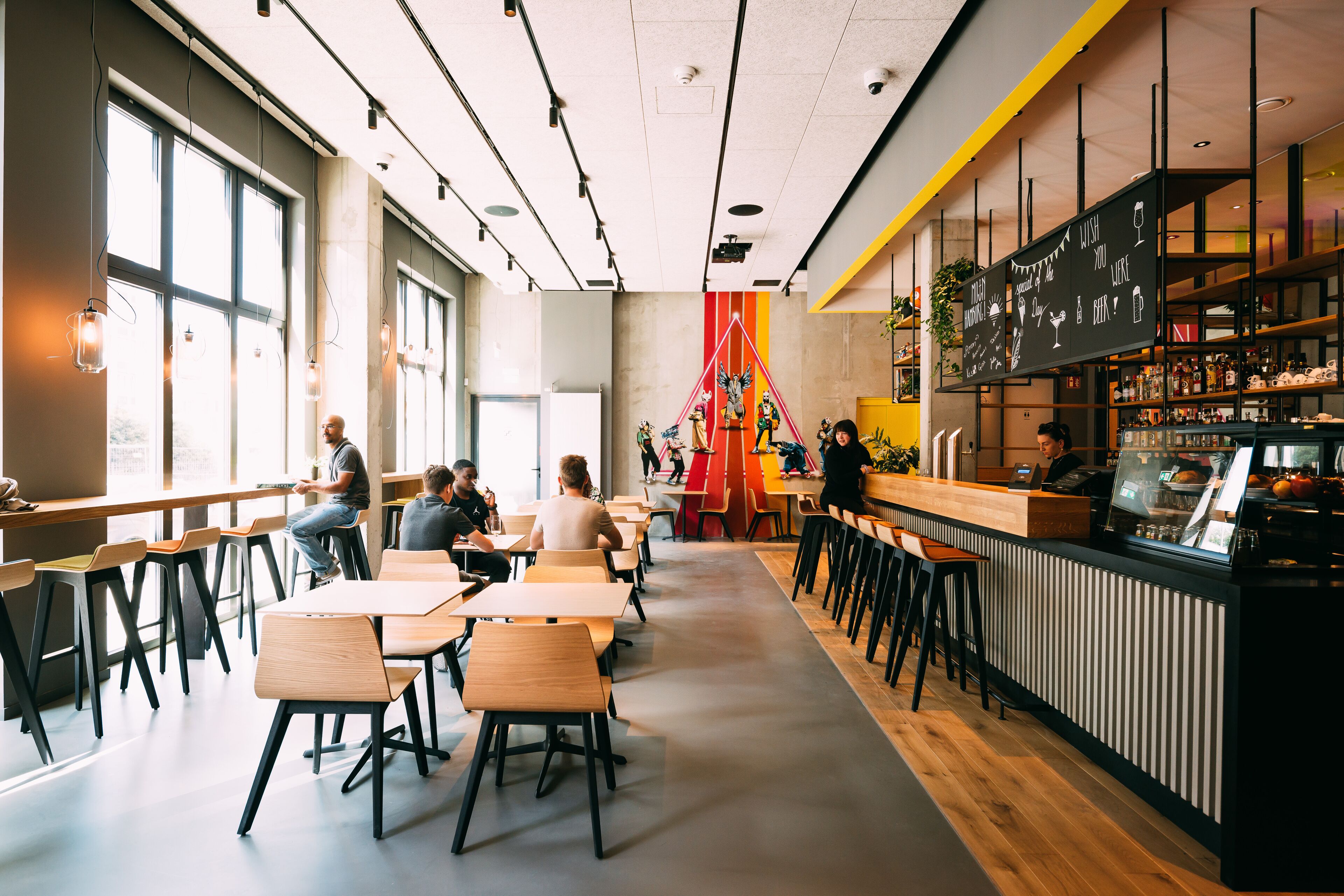 Una cafetería moderna con mesas y sillas de madera, una zona de bar y ventanas amplias que dejan entrar luz natural.