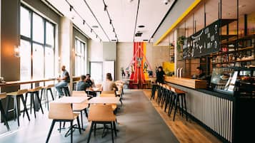 Una cafetería moderna con mesas y sillas de madera, una zona de bar y ventanas amplias que dejan entrar luz natural.