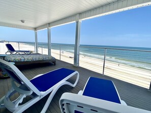 Une terrasse couverte avec deux chaises longues et un banc rembourré, donnant sur une plage et agrémentée d’une rambarde.