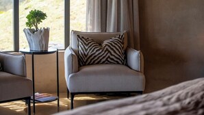 Una sala de estar moderna con un sofá, un sillón con cojines estampados, una mesita auxiliar con una planta y vistas a una ladera a través de amplios ventanales.