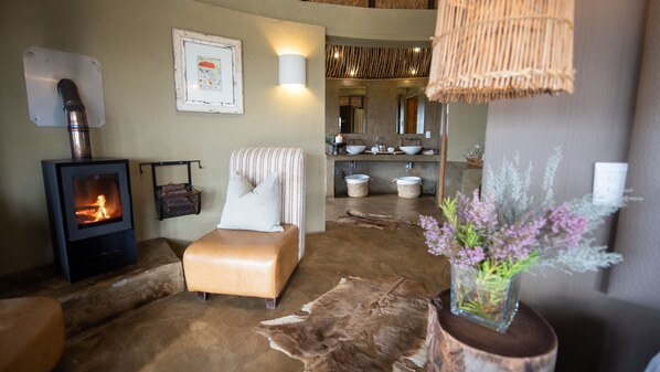 A cozy room with a fireplace, a comfortable chair, and a rustic decor including a cowhide rug and a vase with flowers. - Gondwana Game Reserve (Mossel Bay)