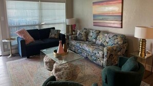 A living room with a dark sofa, a patterned sofa, a glass coffee table, and a green armchair. - Beach Haven - Unit 1303- Located One Block from the Beach! (Port Aransas)