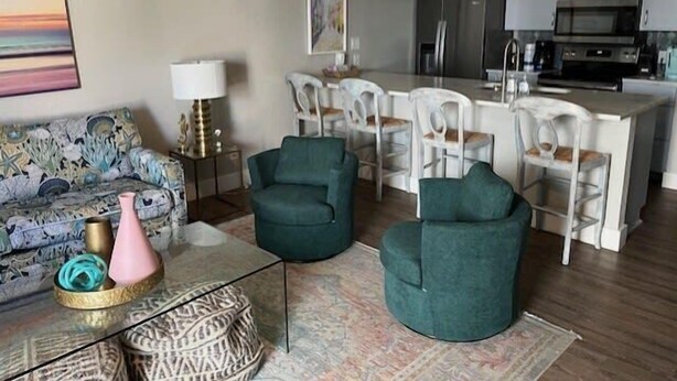 A modern living room with a sofa, two armchairs, a coffee table, and a rug. There is a kitchen area with a refrigerator, microwave, and dining chairs. - Beach Haven - Unit 1303- Located One Block from the Beach! (Port Aransas)