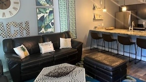 A modern living room with a black leather sofa, a patterned ottoman, and a large clock on the wall. - Island Dunes Condo's-#1307-One block from beach (Port Aransas)